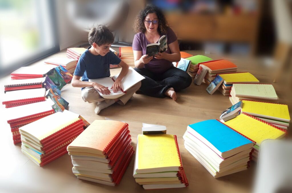 Photo d'un garçon d'une dizaine d'années et d'une dame souriant complices, côte à côte assis en tailleur, au centre d'un grand cercle fait avec de piles de livres en braille et avec le même livre en noir appuyé sur chaque pile de livres en braille. La dame tient entre ses mains un livre en noir et l'enfant tient entre ses mais le même livre en braille papier.