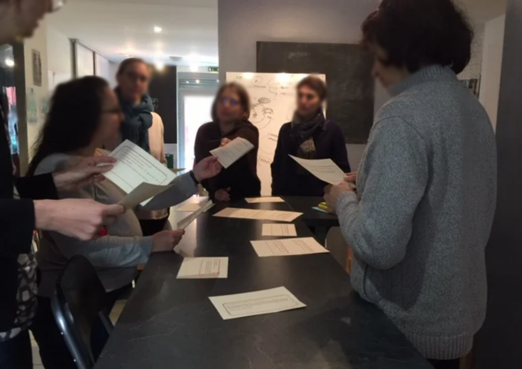 un groupe de 6 personnes est rassemblé debout autour d’une table rectangulaire. Ils tiennent des brochures dans les mains et semblent écouter attentivement l’une des personnes du groupe.