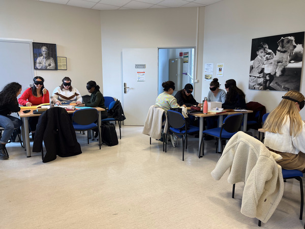 Dans une salle de cours, trois groupes de femmes sont assises autour des trois tables rectangulaires. Elles portent des bandeaux occultant sur leurs yeux et sont concentrées à la découverte tactile de différentes activités.